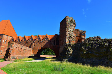 Old brick dilapidated wall, arched passage under the path and a tower with a red roof. Ruins of the fortress. Ancient building against a blue sky background and green lawn. Poland, Torun, August 2023.