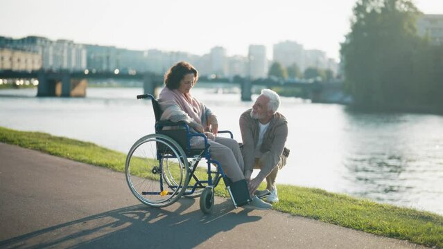 Husband walking with wife in wheelchair. Senior family couple spending time in city park near pond. Woman hugging man showing love care, they smiling. Living with physical disability concept.