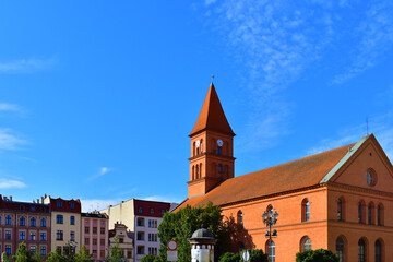 Ancient catholic church with tower, clock, red roof, made of red brick. Ancient architecture. Green trees and other buildings around, blue sky. Torun, Poland, August 2023 