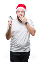 Young caucasian man wearing christmas hat holding credit card over isolated background cover mouth with hand shocked with shame for mistake, expression of fear, scared in silence, secret concept