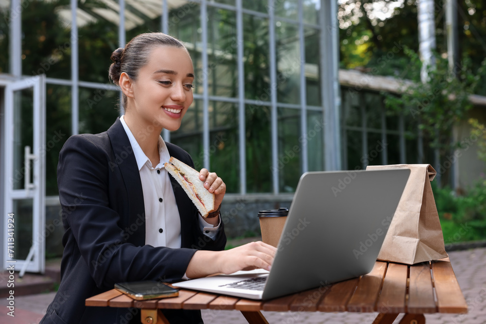 Wall mural Happy businesswoman with sandwich using laptop while having lunch at wooden table outdoors