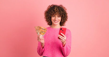 Excited woman with cash and looking mobile phone in pink studio - obrazy, fototapety, plakaty