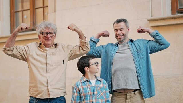 People standing in front of yellow wall and pose to camera how strong they are. Caucasian grandfather, son and grandkid looking at camera and smiling. Family reunion. Funny ideas for picture.