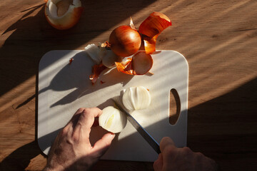 Male hands slicing onion with a knife on a kitchen table, on white cutting board, with sunlight from window, top view