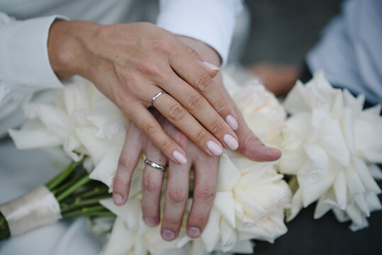 Wedding rings on the hands of newlyweds. Cropped image of bride and groom, little fingers with rings on a bouquet of white flowers background. Concept of promise. Vow. Engagement.