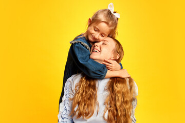 Little caring girl, kid hugging her smiling happy mother against yellow studio background. Showing...