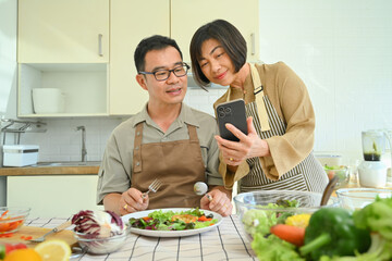 Smiling senior couple using smart mobile phone together while cooking in kitchen.