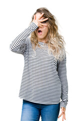 Beautiful young blonde woman wearing stripes sweater over isolated background peeking in shock covering face and eyes with hand, looking through fingers with embarrassed expression.