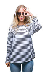 Beautiful young blonde woman wearing headphones and sunglasses over isolated background doing ok gesture with hand smiling, eye looking through fingers with happy face.