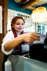 Vertical photo of a dark-skinned waitress working in uniform in a coffee shop. An African woman with braided hair is pulling out a bill on the restaurant's touch computer.Woman working in hospitality.