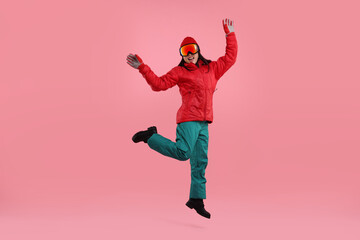 Fototapeta na wymiar Happy woman in winter sportswear and goggles jumping on pink background