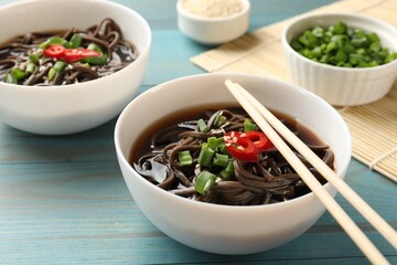 Tasty soup with buckwheat noodles (soba), chili pepper, green onion in bowl and chopsticks on light blue wooden table, closeup