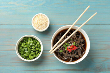 Tasty buckwheat noodle (soba) soup with chili pepper, green onion, sesame and chopsticks on light blue wooden table, flat lay - obrazy, fototapety, plakaty