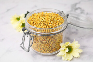 Fresh bee pollen granules in jar and flowers on light table, closeup