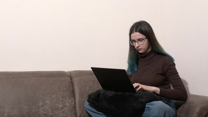 A serious young woman using a laptop, sitting at home on the couch, typing, shopping or chatting online on a social network, working on a project, an exam. Online education.