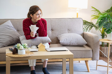 A young Asian woman sits on a sofa in her home, feeling worried and frustrated about her monthly expenses. Various utility bills.