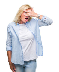 Middle age blonde woman over isolated background peeking in shock covering face and eyes with hand, looking through fingers with embarrassed expression.