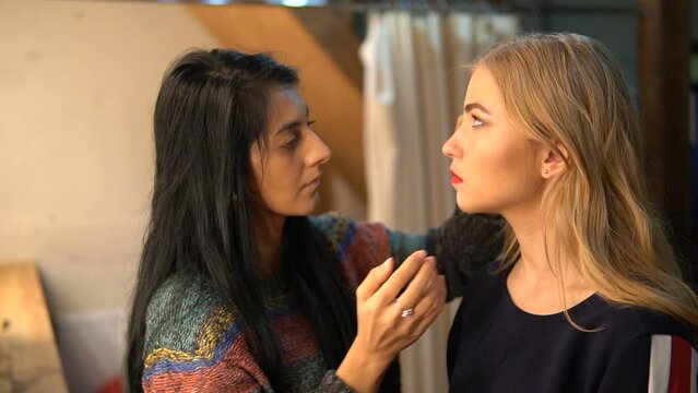 A young actress is doing makeup. The make-up artist applies make-up on the face of a beautiful girl. The model is preparing for shooting in the make-up room. A blond woman in front of a mirror.