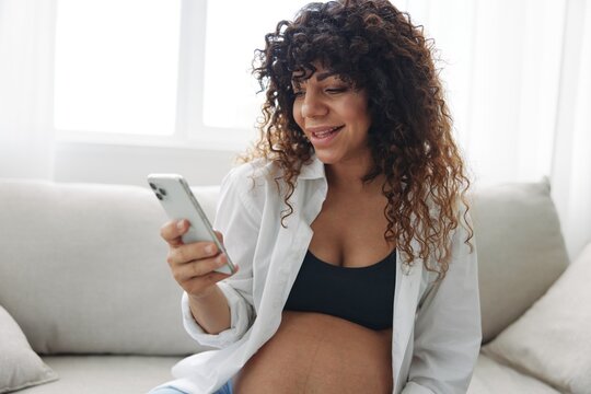 Pregnant woman blogger sits on the couch at home and takes pictures of herself on the phone, selfie and video call, online doctor consultation, pregnancy management, freelance work smiling