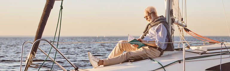 Relaxed senior man reading a book, while sitting on the side of sail boat or yacht deck floating in...