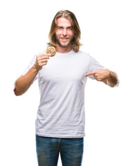 Young handsome man with long hair eating chocolate cooky over isolated background with surprise face pointing finger to himself