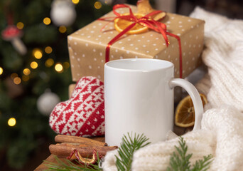 Fototapeta na wymiar White coffee mug near Christmas present, cosy heart and sweater, winter mockup