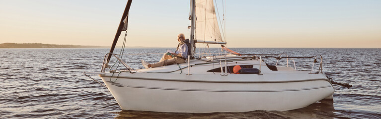 Luxury lifestyle. View of the sail boat or yacht floating in sea with relaxed senior man reading a book, sitting on the deck