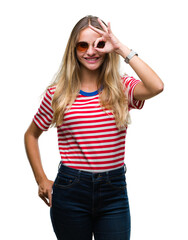 Young beautiful blonde woman wearing sunglasses over isolated background doing ok gesture with hand smiling, eye looking through fingers with happy face.