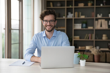 Millennial office employee in glasses sitting at desk with laptop smiling looking at camera....