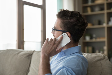 Close up side view man talks on smartphone rest on sofa. Young guy in glasses relaxing alone at home looking into distance lead personal conversation, make order remotely. Modern tech, communication