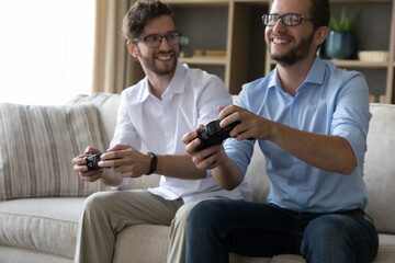 Two millennial male twins relax on weekend together sit on sofa at home holding gamepads, play console videogames enjoy leisure activity and joint hobby, having friendly relationships. Family pastime