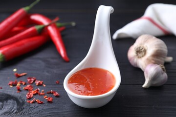 Spicy chili sauce in spoon, peppers and garlic on black wooden table, closeup