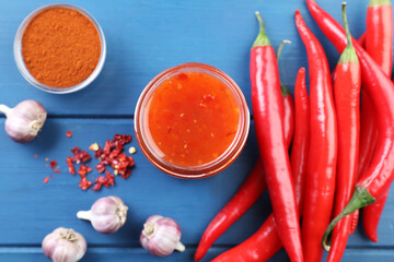 Spicy chili sauce in jar and ingredients on blue wooden table, flat lay