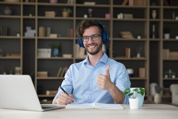 Young man in glasses wear headphones sit at desk with laptop smile look at camera showing thumb up...