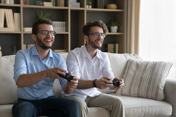 Two excited young adult brothers spend time sit on sofa with joypads in hands, play videogames,...