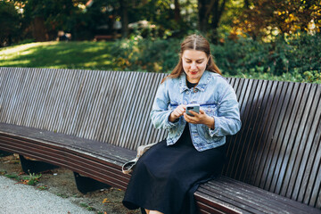 Obraz premium Positive cheerful woman tourist dressed in casual look holding smartphone gadget in hand for communicate on the city street. Happy woman chatting on cell smartphone and relaxing on the bench outdoors