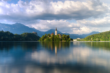 Serene waters of Lake Bled reflect the idyllic church island and castle amidst the Julian Alps on a cloudy day