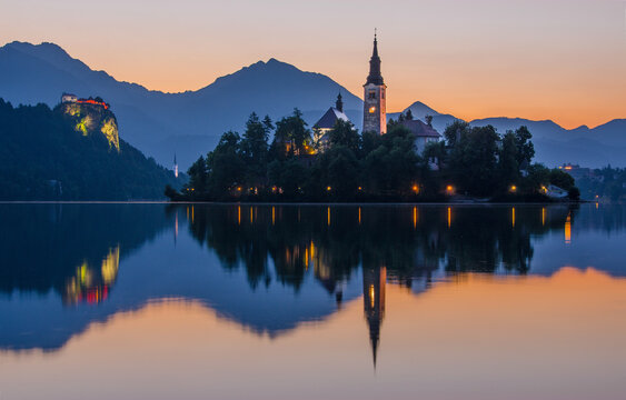 The sun sets on Lake Bled, casting a warm glow on the Pilgrimage Church of the Assumption of Maria and the Bled Castle