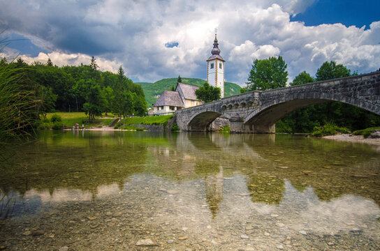 A stone bridge arches over the crystal-clear waters of Lake Bohinj with the Church of St. John the Baptist and mountains in the backdrop