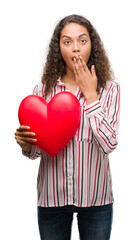 Fototapeta na wymiar Young hispanic woman in love holding red heart cover mouth with hand shocked with shame for mistake, expression of fear, scared in silence, secret concept