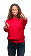 Young hispanic woman wearing red sweater approving doing positive gesture with hand, thumbs up smiling and happy for success. Looking at the camera, winner gesture.