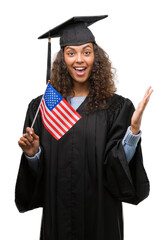 Young hispanic woman wearing graduation uniform holding flag of United States very happy and excited, winner expression celebrating victory screaming with big smile and raised hands