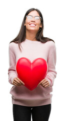 Young hispanic woman holding heart in love with a happy face standing and smiling with a confident smile showing teeth