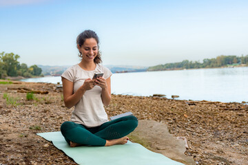 Fototapeta na wymiar In between exercises, she uses her phone to remind herself of the next moves while sitting on a workout mat by the river on the beach.