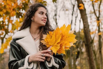 Attractive pretty woman walking outdoor, portrait of young lady in warm sunny autumn park season, fall, hold yellow orange red maple leaves, dressed leather wool fur jacket smiling having fun 