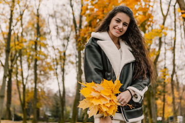 Attractive pretty woman walking outdoor, portrait of young lady in warm sunny autumn park season, fall, hold yellow orange red maple leaves, dressed leather wool fur jacket smiling having fun 