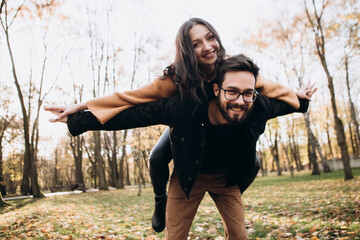 Heterosexual caucasian young loving couple, husband carrying on back his wife hands wings fly, hugging smiling kissing laughing spending time together. Autumn, fall season, orange yellow red maple lea - obrazy, fototapety, plakaty
