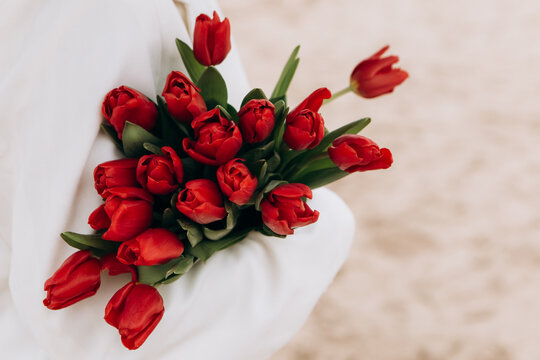 Attractive brunette woman walking on the beach shore in moody cloudy windy weather with bouquet of red tulips flowers, dressed in white suit jacket. International Women`s Day 8th March concept - Powered by Adobe
