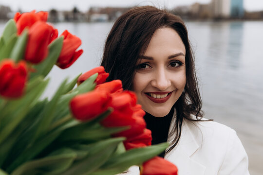 Attractive brunette woman walking on the beach shore in moody cloudy windy weather with bouquet of red tulips flowers, dressed in white suit jacket. International Women`s Day 8th March concept