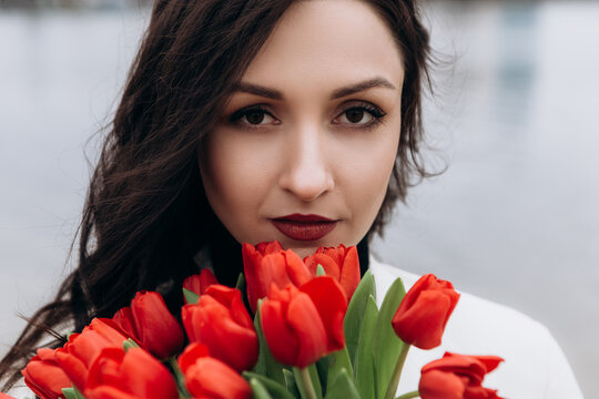 Attractive brunette woman walking on the beach shore in moody cloudy windy weather with bouquet of red tulips flowers, dressed in white suit jacket. International Women`s Day 8th March concept
